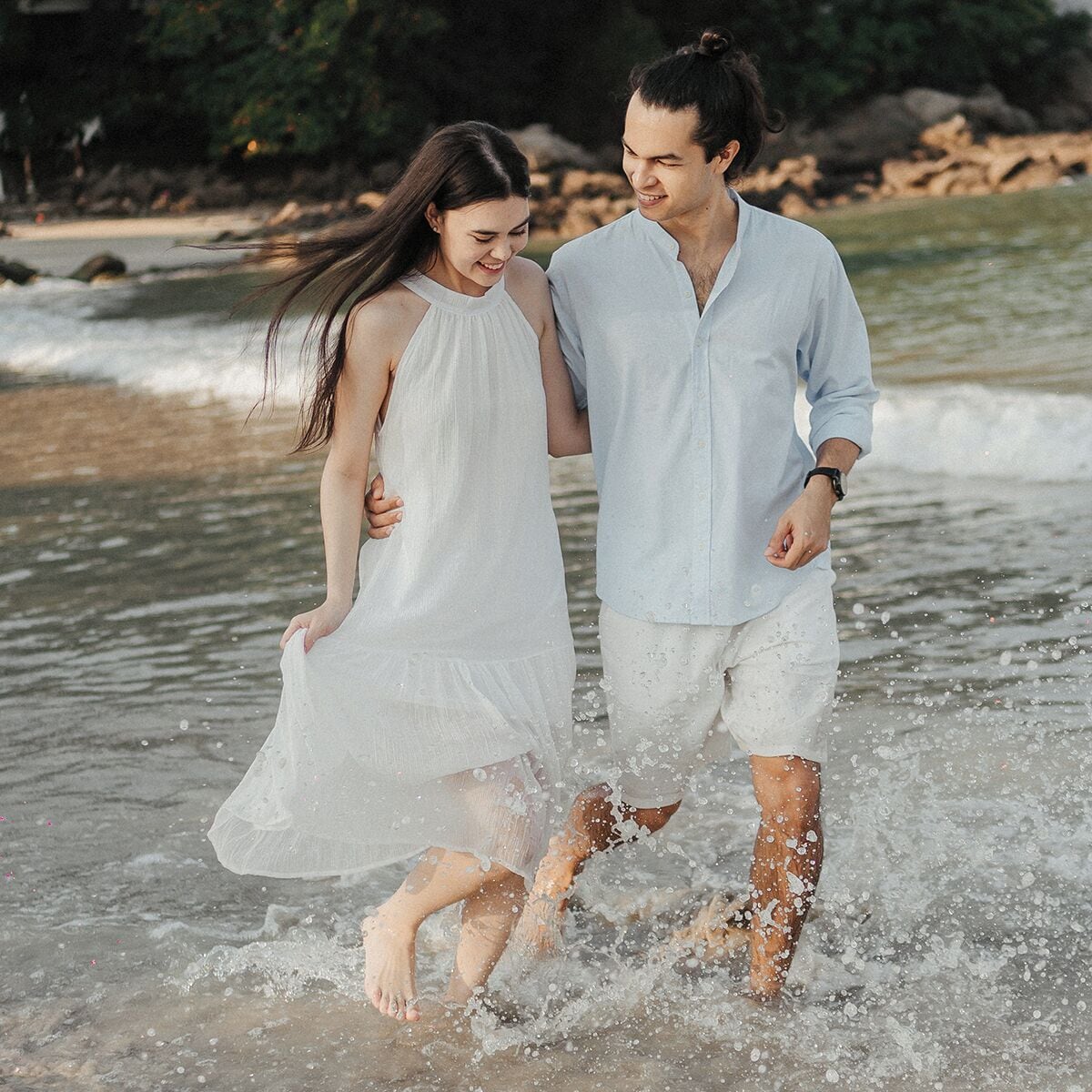 Una pareja caminando por aguas poco profundas en una playa con una costa rocosa al fondo.