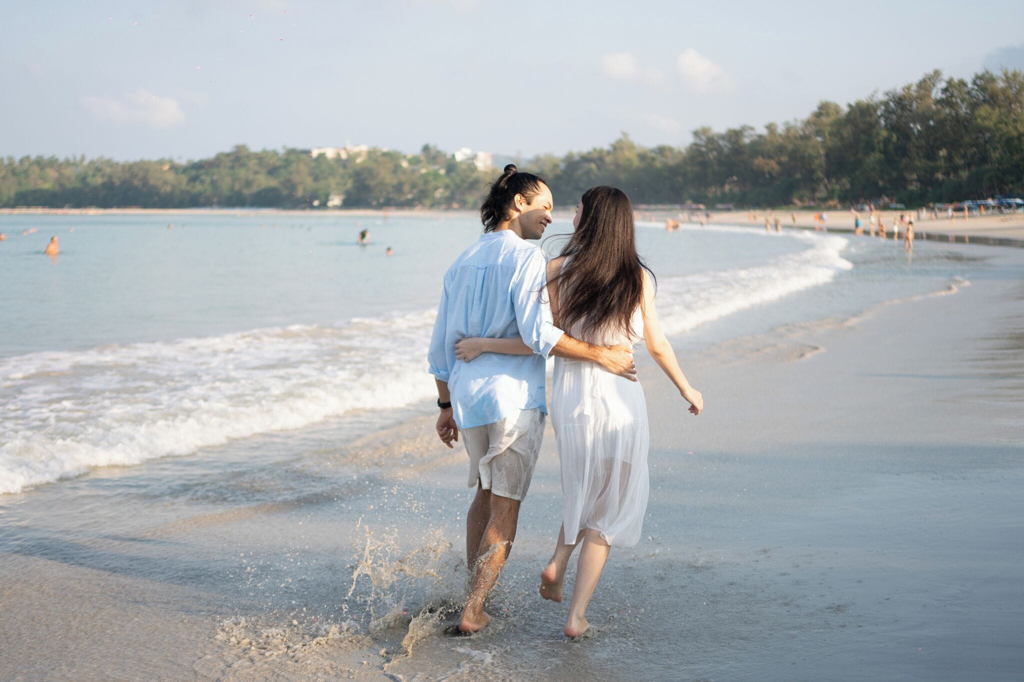 Una pareja caminando en una playa con olas y una costa lejana.