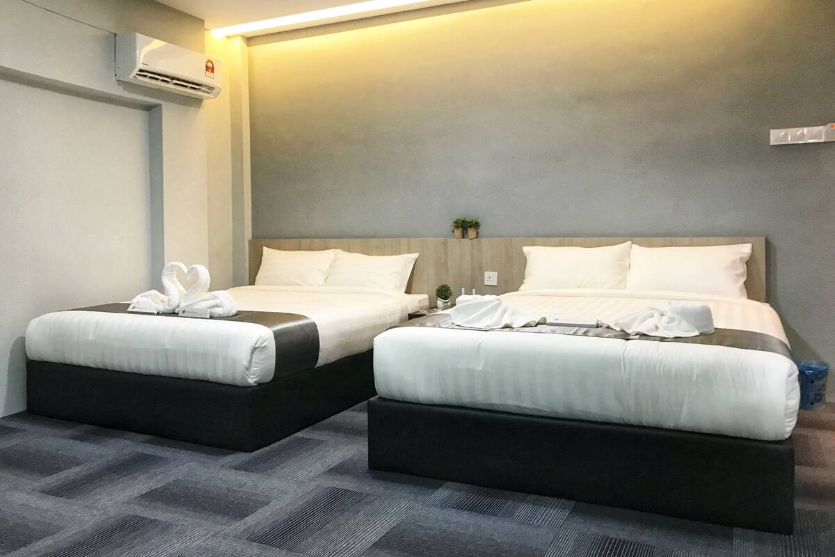 A hotel room with two beds, each with white linens and a heart-shaped towel arrangement. There is a wall-mounted air conditioner and a small plant on the nightstand.