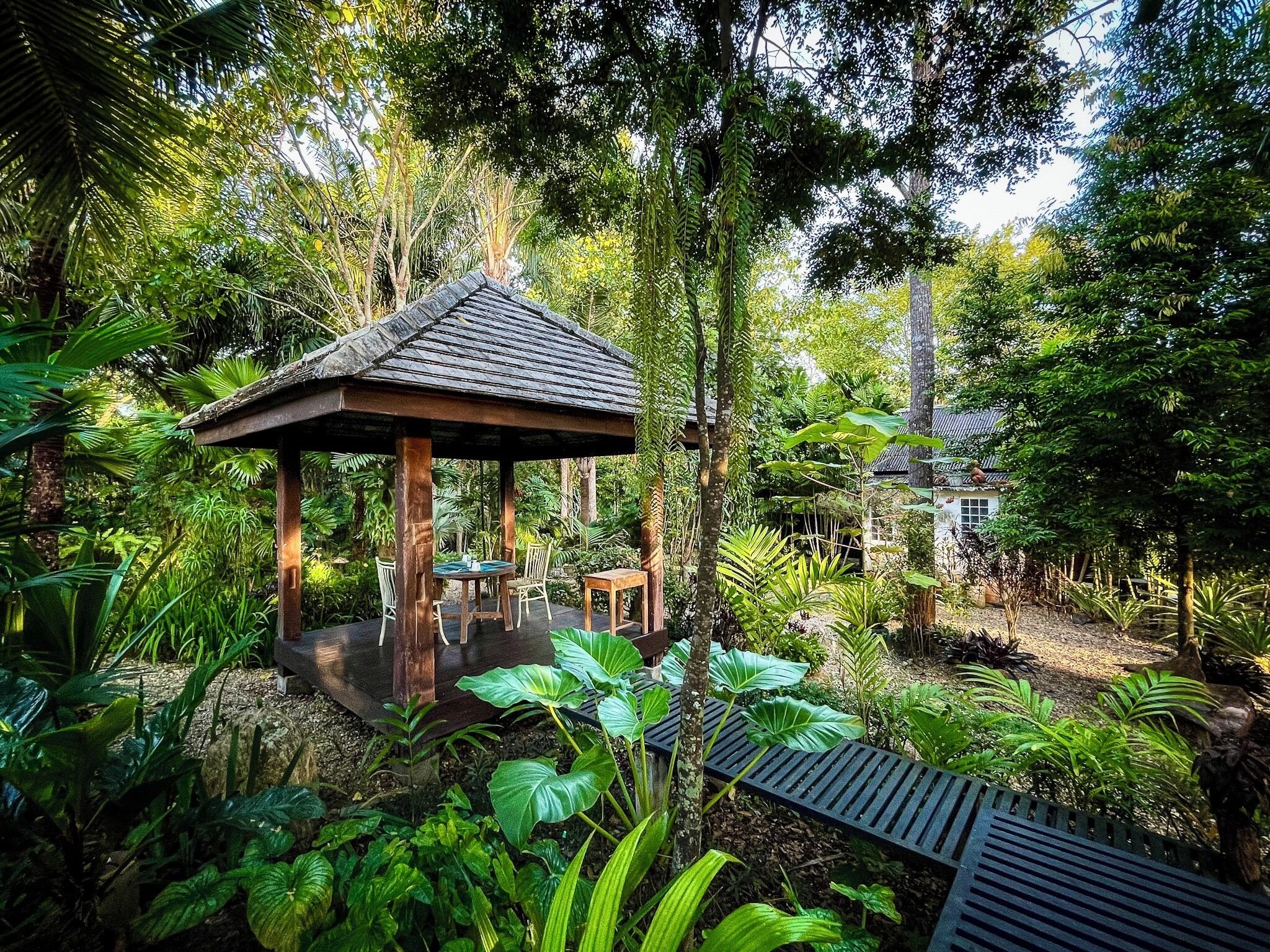 A wooden gazebo with a tiled roof surrounded by lush greenery and a small table with chairs.