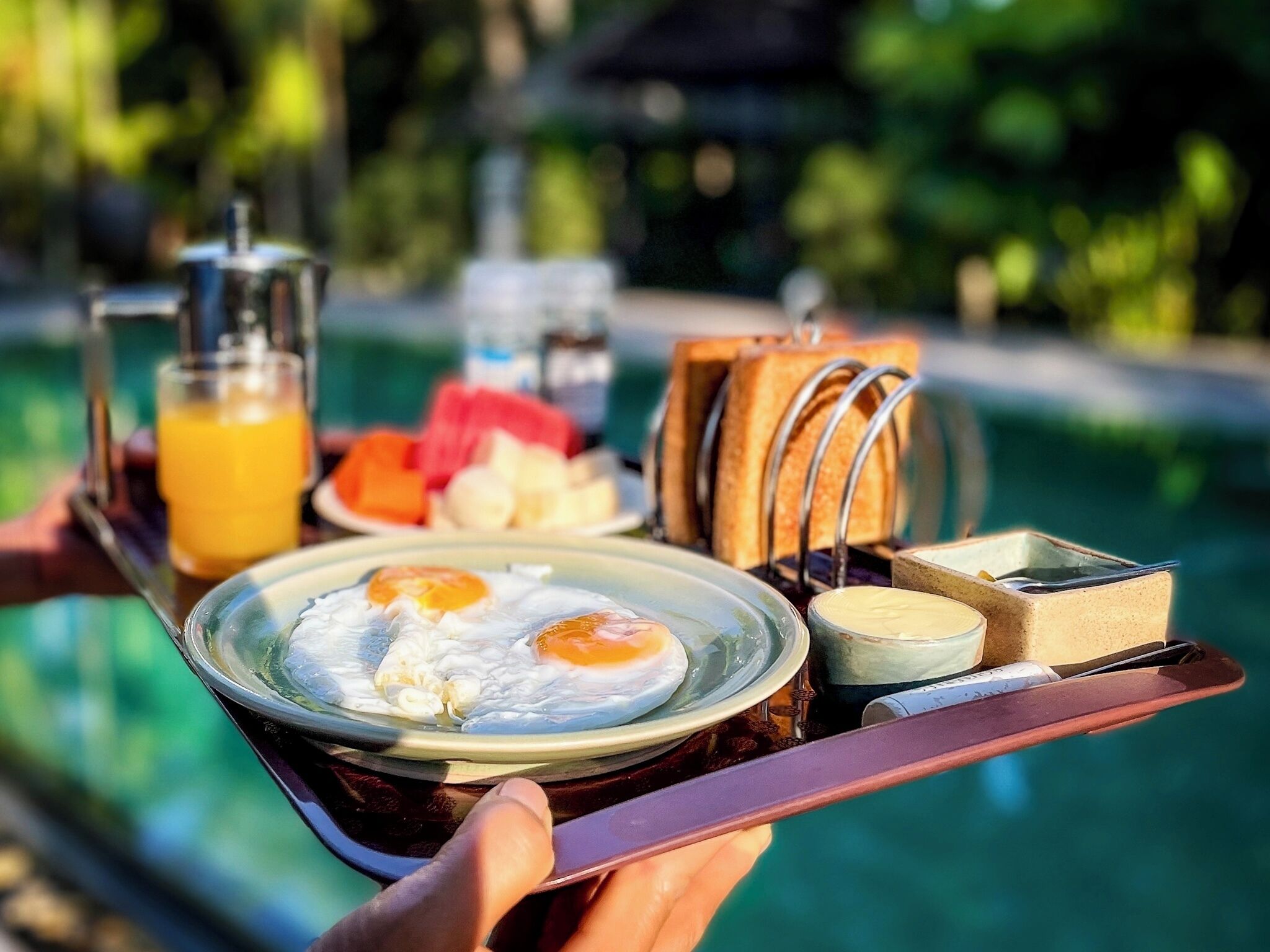 A breakfast tray with eggs, toast, fruit, and juice by a pool.