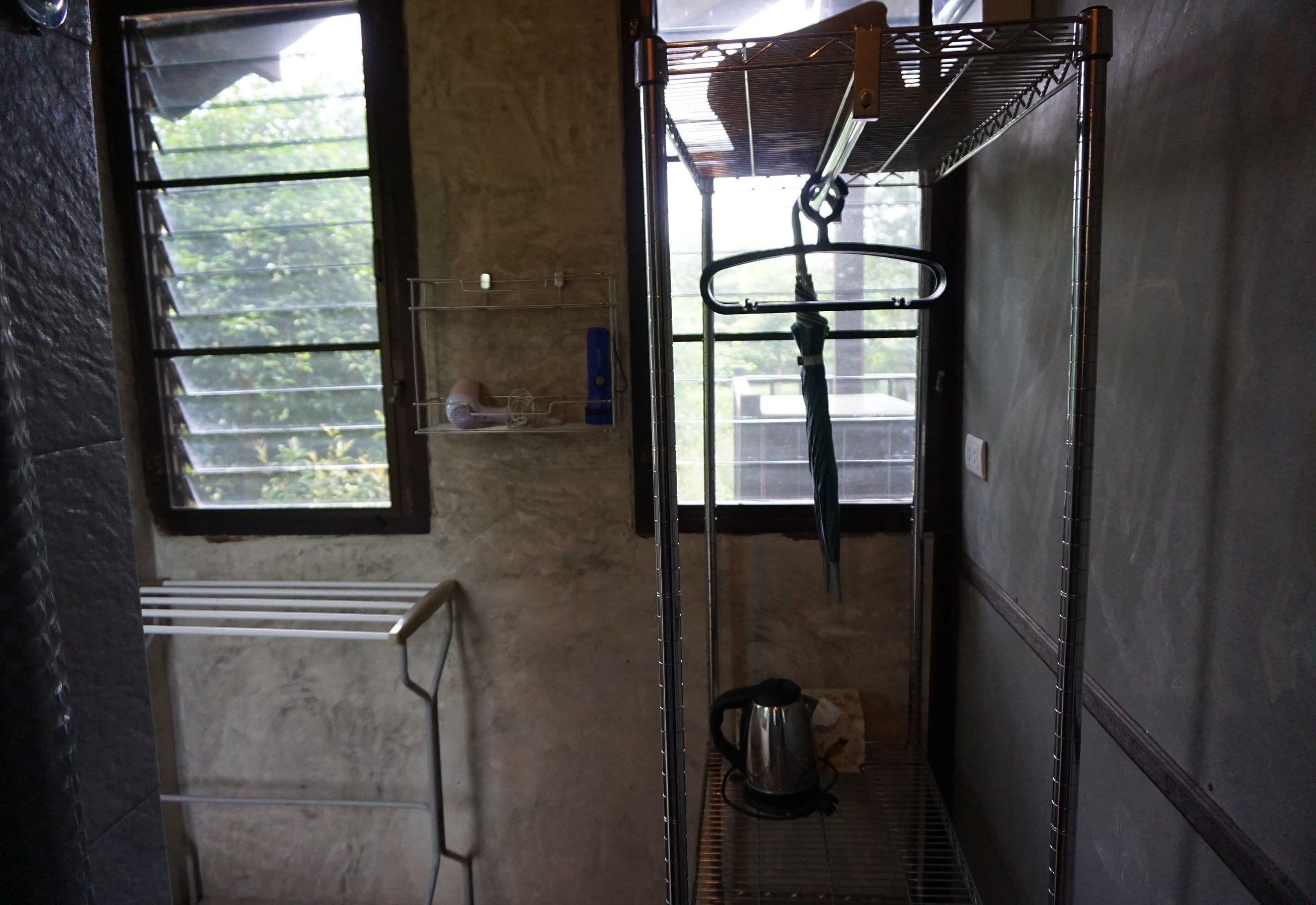 A narrow corridor with a metal rack, a kettle, and a window with a view of greenery.