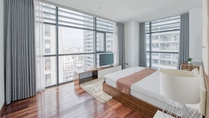 A modern hotel room with a large bed, a TV, and a view of the city through floor-to-ceiling windows.