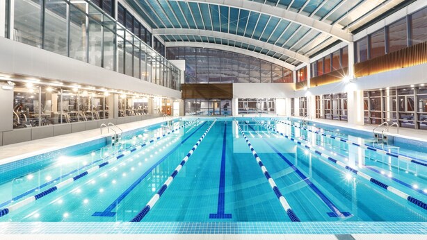 A large indoor swimming pool with multiple lanes, surrounded by glass walls and a high ceiling with skylights.