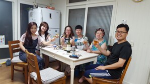 A group of people sitting around a table with food and drinks, in a room with a refrigerator and a window.