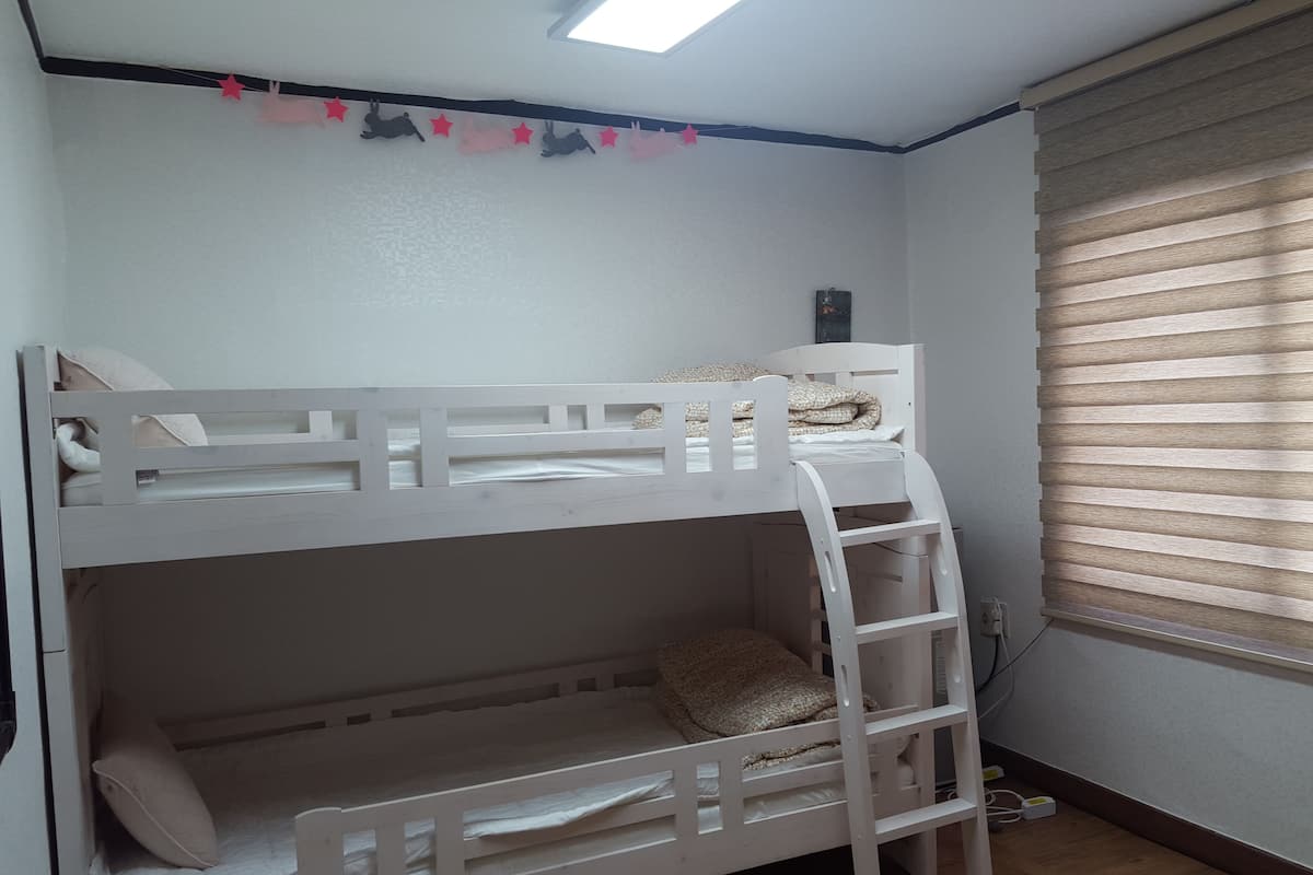 Een slaapkamer met een stapelbed, raam en zonwering, een wandplank en een stopcontact.
