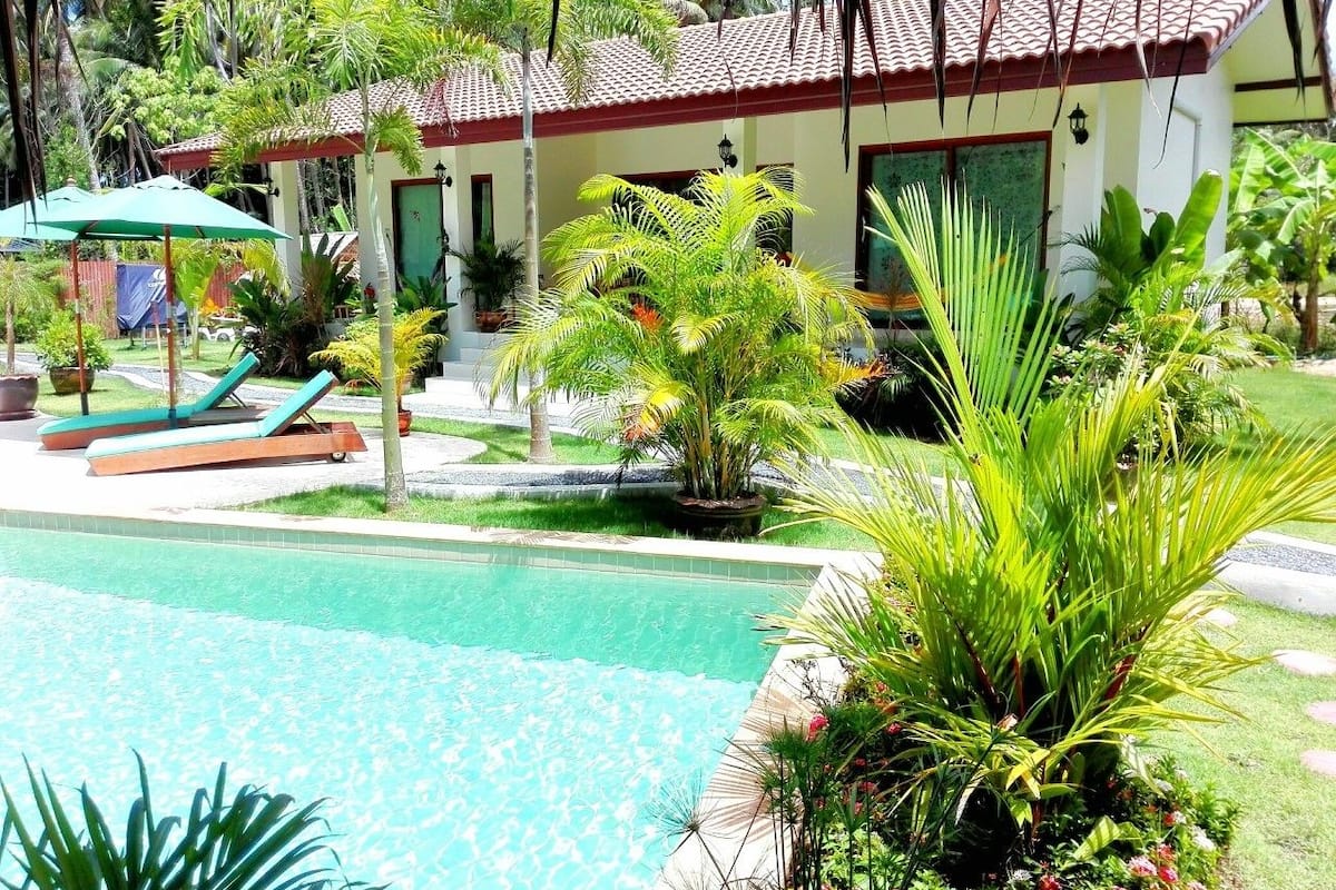 Un área de piscina con sillas de descanso, un edificio pequeño con techo de tejas y diversas plantas tropicales.