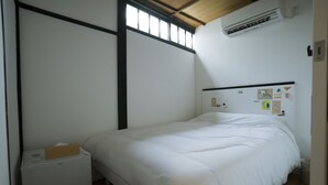 Ein Zimmer mit einem Einzelbett, Holzboden, einem weißen Bett, einem kleinen Nachttisch, einer Wand-Klimaanlage und einem Fenster mit Jalousien.