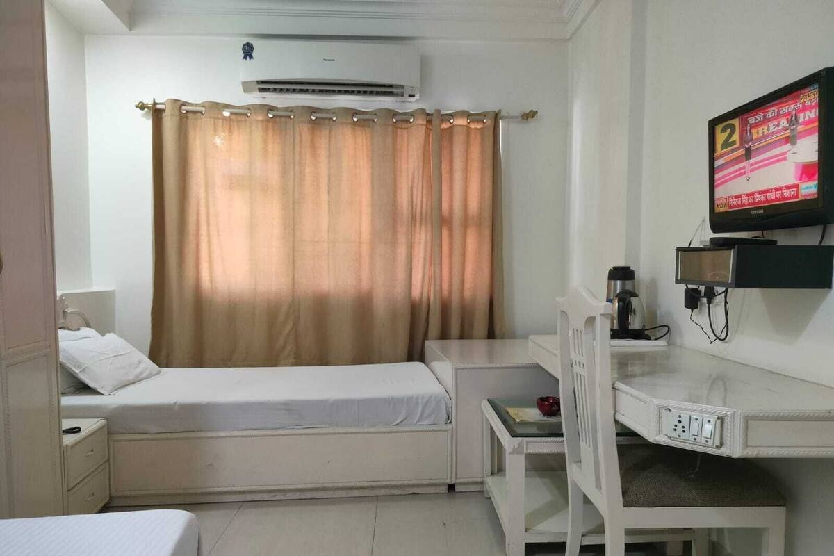 A small, clean room with a single bed, a desk with a chair, a television, and an air conditioning unit.