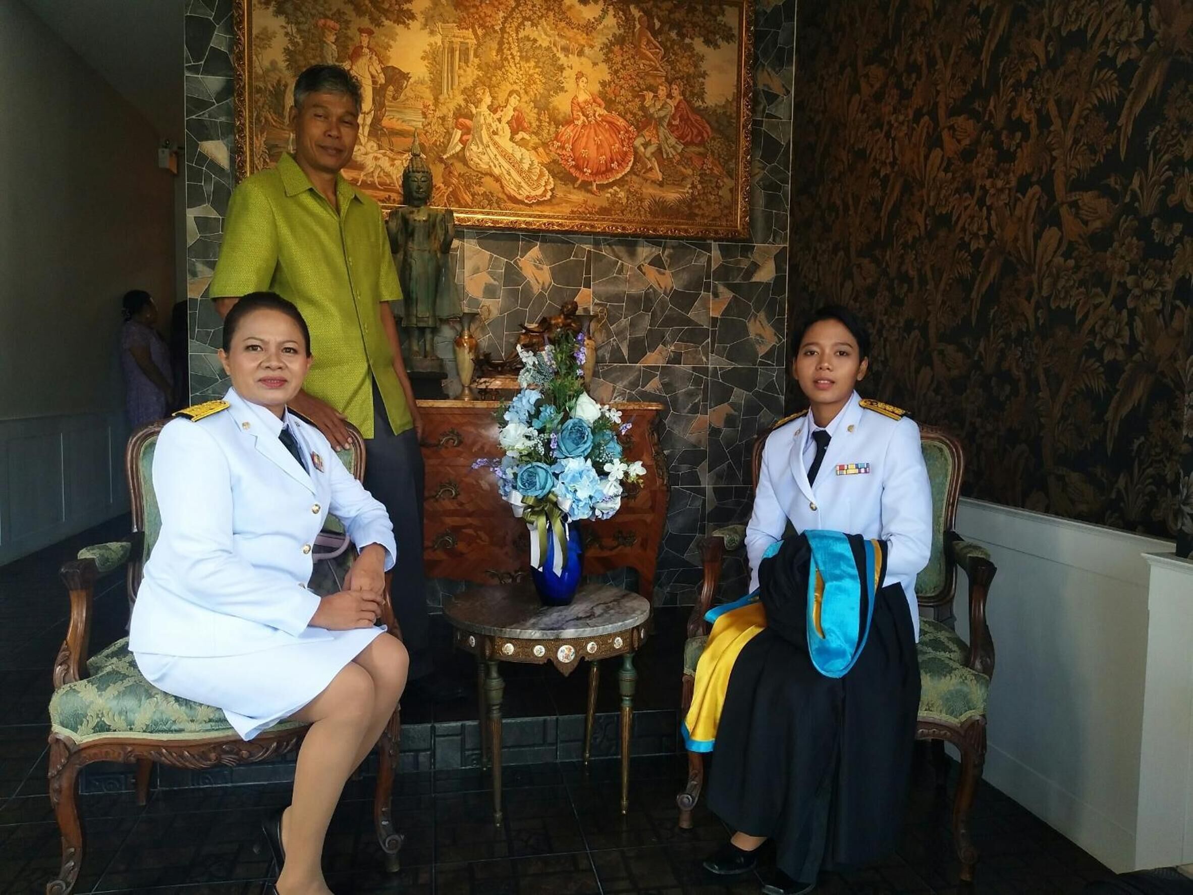 Una sala elegante con dos personas sentadas en sillones decorados, una mesa con un jarrón de flores y un mural en la pared.