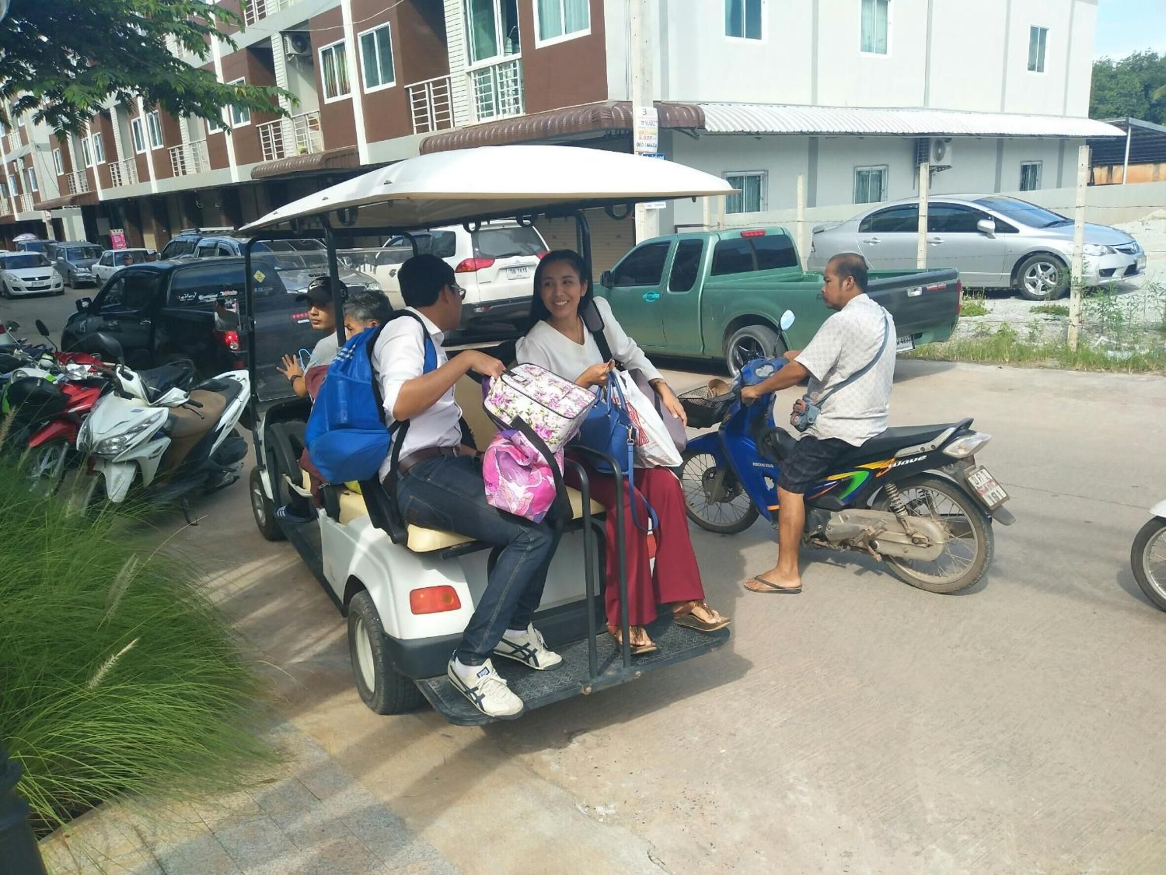 A group of people on a motorized cart with a canopy, parked in a lot with cars and motorcycles.