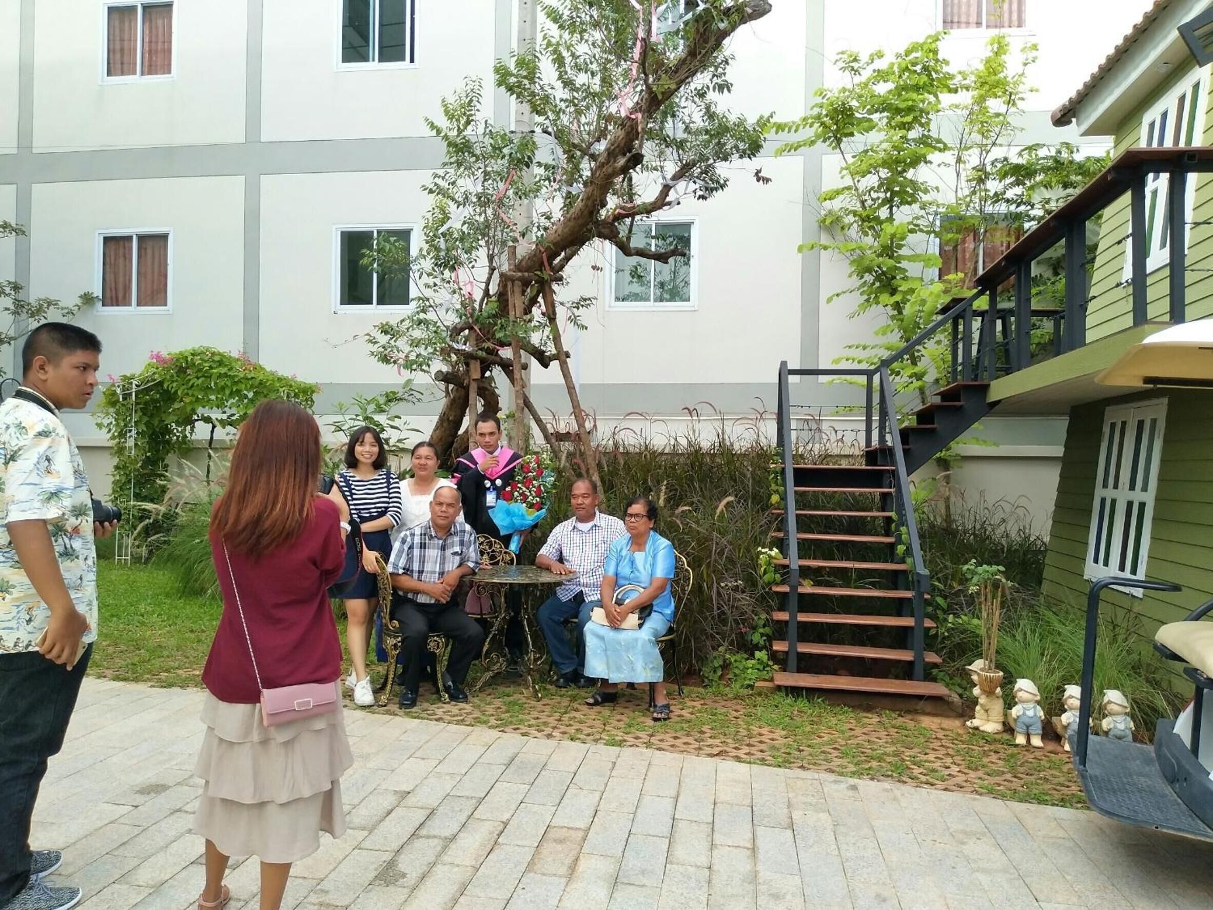 A group of people seated around a table in a courtyard with a staircase and greenery.