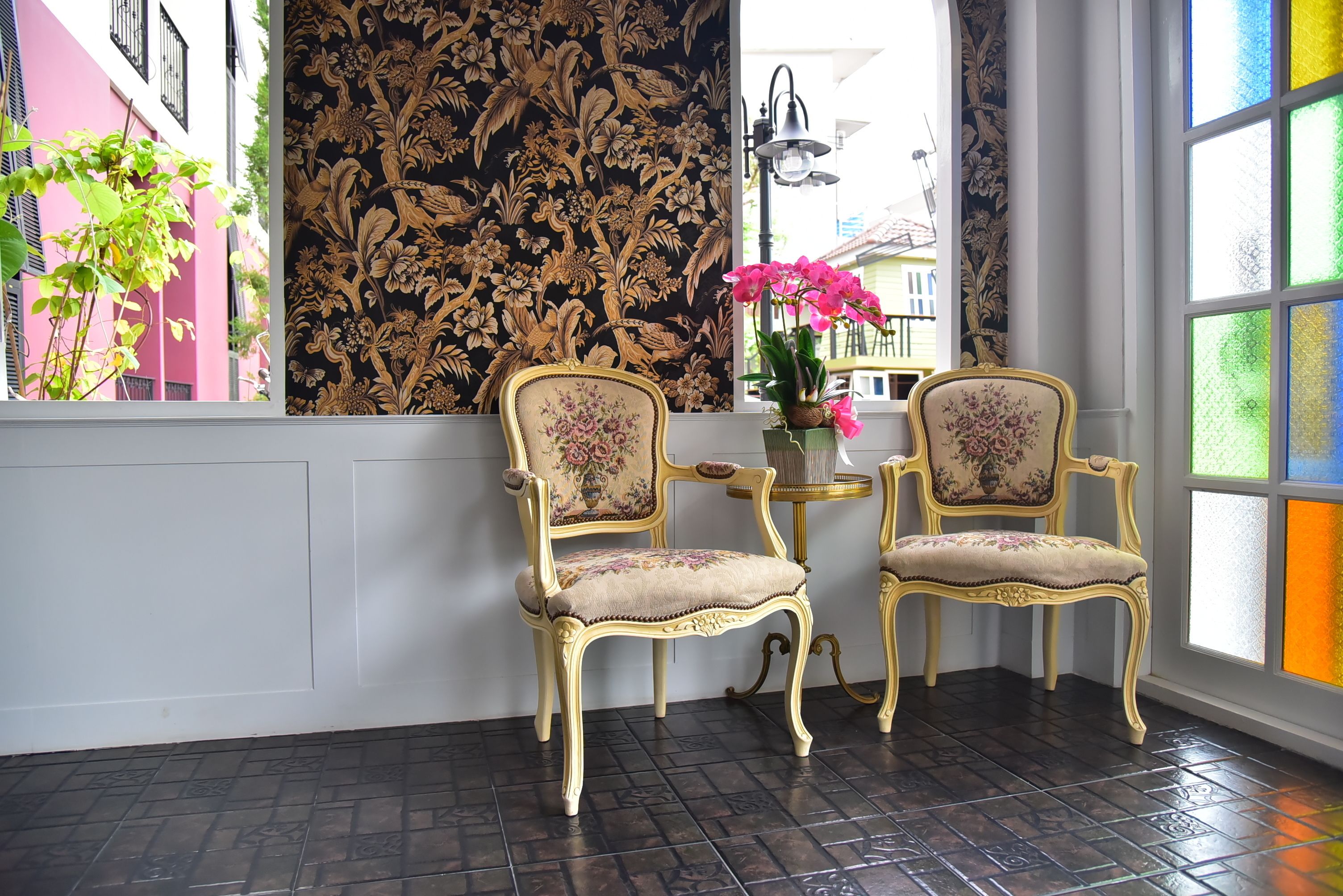 Una habitación con dos sillas ornamentadas, una mesita con un florero y vistas a edificios coloridos a través de una ventana con cortinas estampadas.