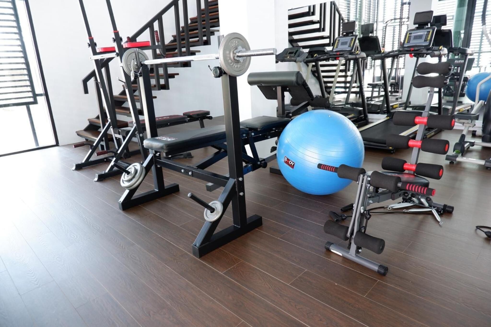 A gym with various exercise equipment including a bench press, a barbell, a blue medicine ball, and dumbbells.
