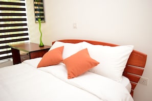 A neatly made bed with white and orange pillows, a green vase on a bedside table, and a window with blinds.