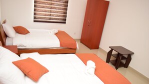 A hotel room with two beds, each with white bedding and orange accents, a wooden wardrobe, and a small wooden side table.