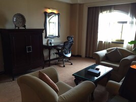 Habitación de hotel con escritorio, una silla de oficina, dos sillones, una mesita de centro, un espejo grande y un plato decorativo en un aparador.