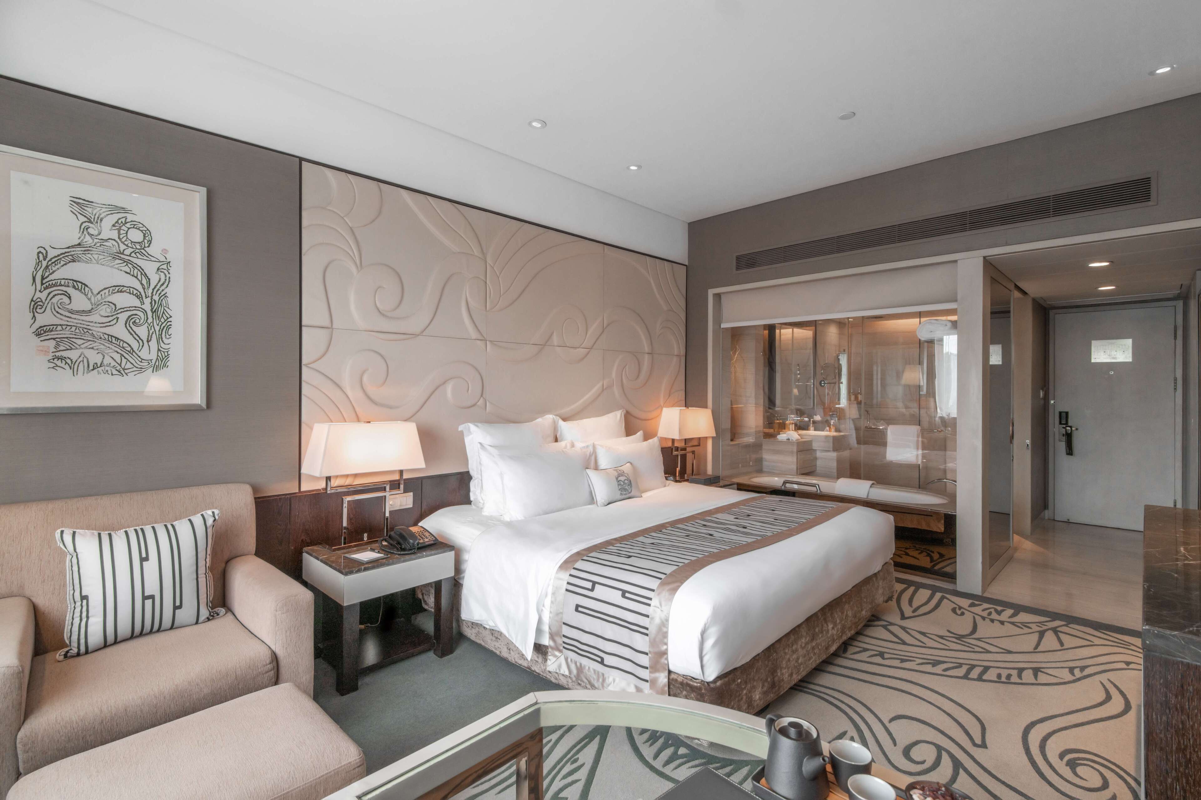 Ein modernes Hotelzimmer mit einem großen Bett, einer Couch, einem Schreibtisch mit Lampe und einem durch eine Glastrennwand einsehbaren Badezimmer.