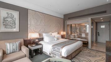 Ein modernes Hotelzimmer mit einem großen Bett, einer Couch, einem Schreibtisch mit Lampe und einem durch eine Glastrennwand einsehbaren Badezimmer.