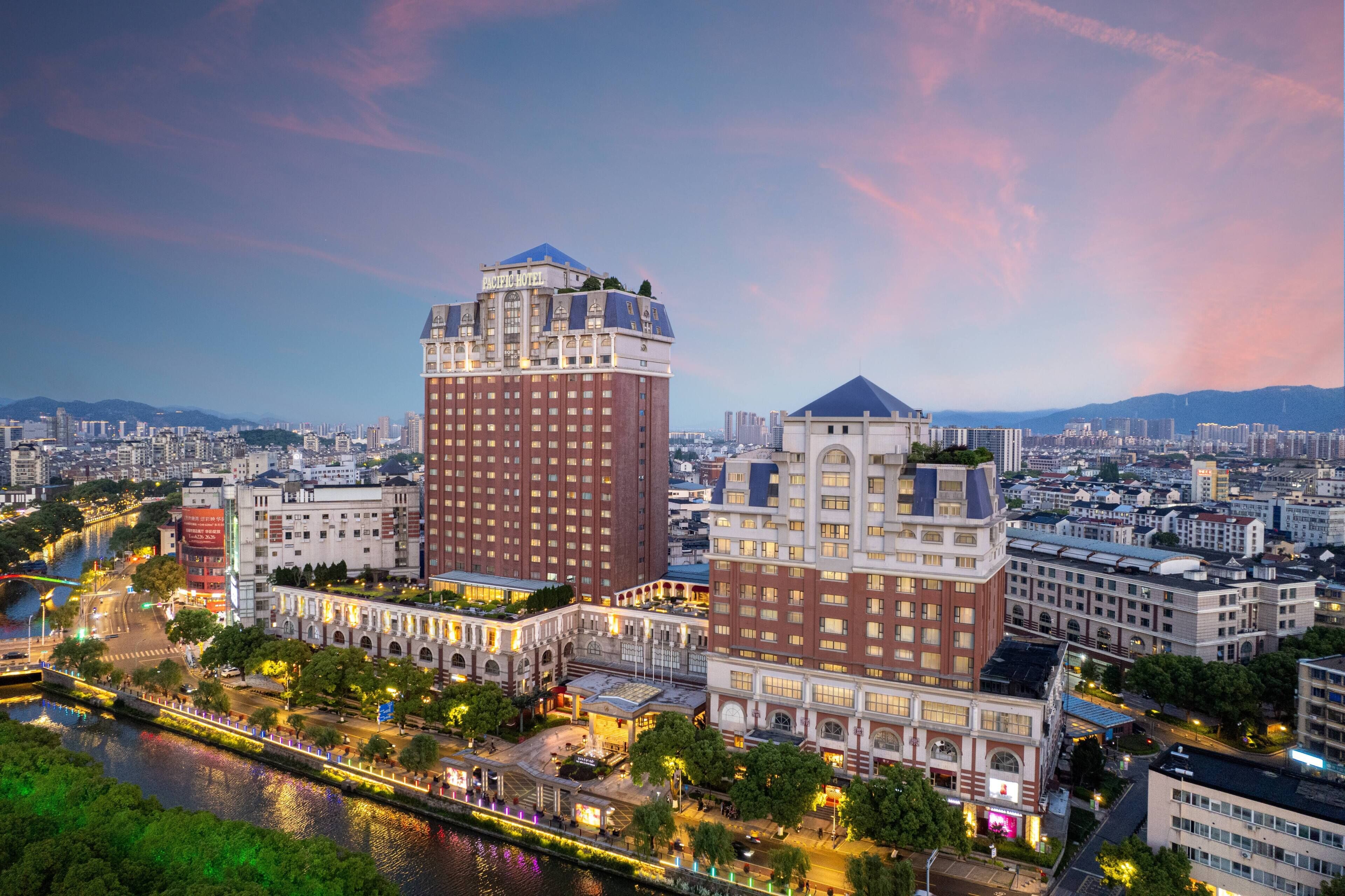 Un paisaje urbano con un hotel destacado de diseño moderno, un edificio histórico con ventanales en arco, y un río que atraviesa el primer plano.