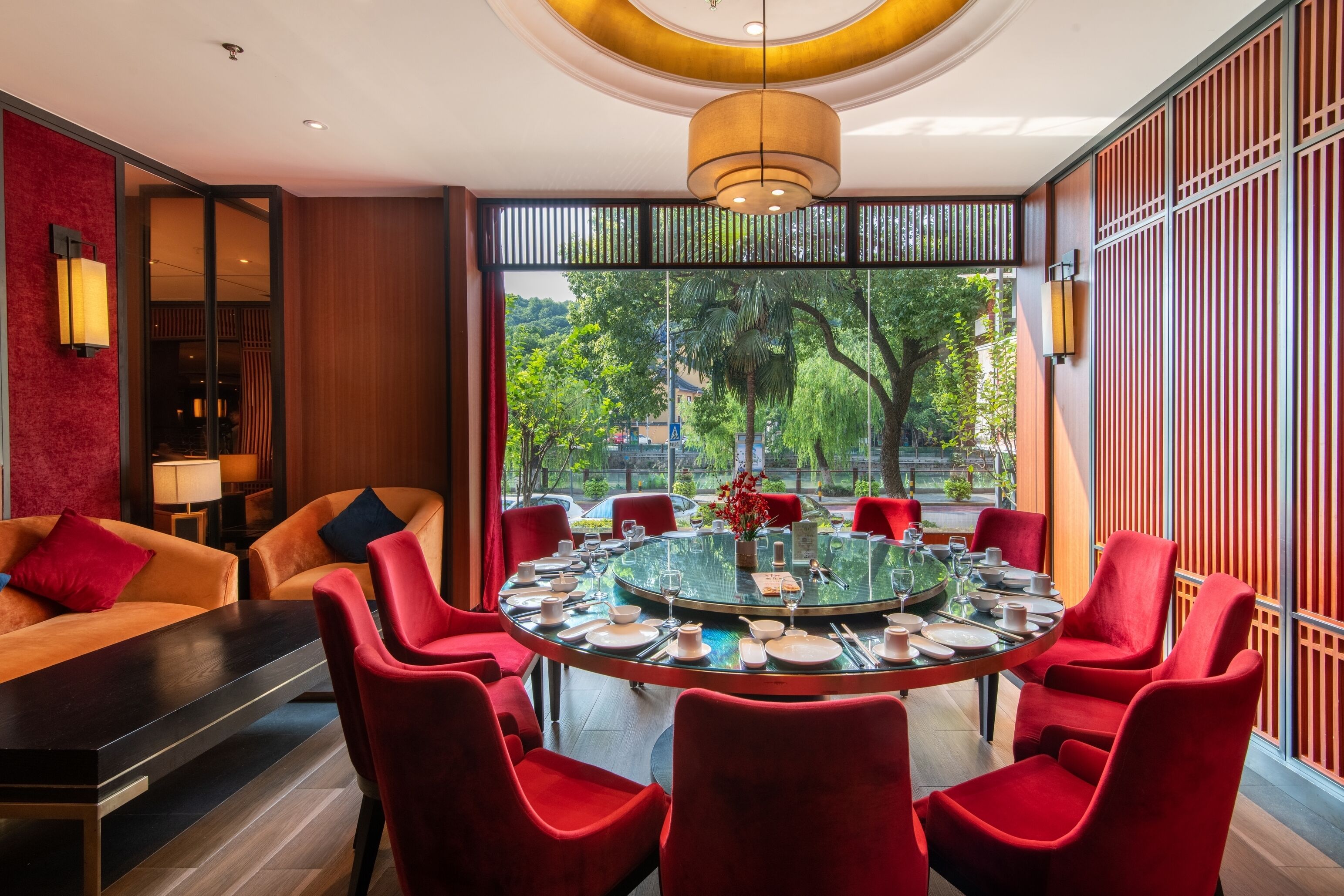 A modern dining area with a round glass table surrounded by red chairs, a sofa, and a red accent wall.