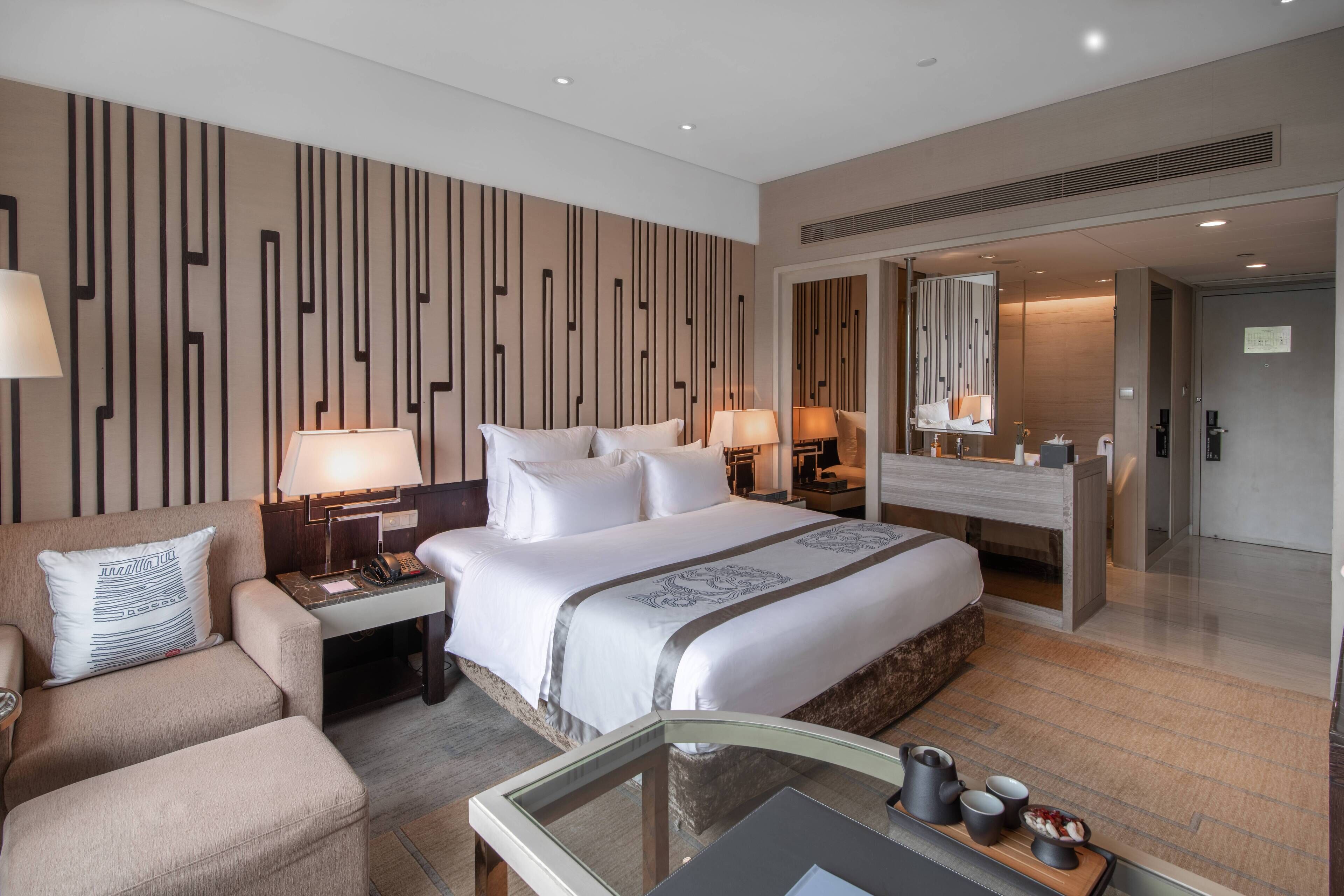 A modern hotel room with a large bed, a sofa, a desk, and a bathroom visible through an open door.