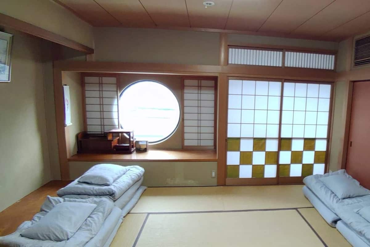 Een traditionele Japanse kamer met tatami vloerbedekking, een futonbed, een shoji scherm en uitzicht door een rond raam.