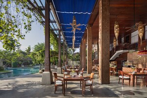 Uma área de refeições ao ar livre com mesas e cadeiras, piscina e galeria coberta.