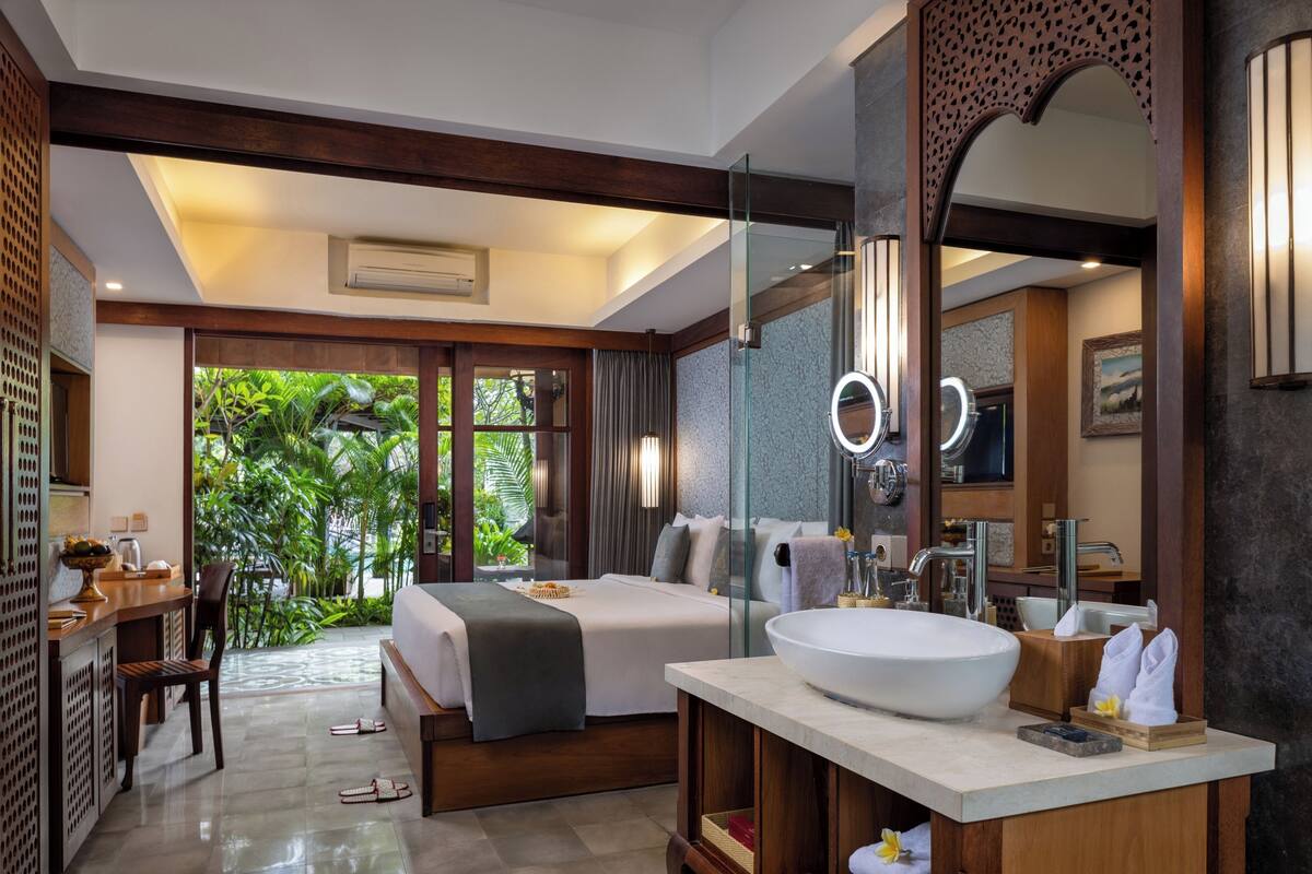A modern hotel room with a large bed, a sitting area with a desk and chair, a large bathroom with a round mirror, and a view of lush greenery outside.
