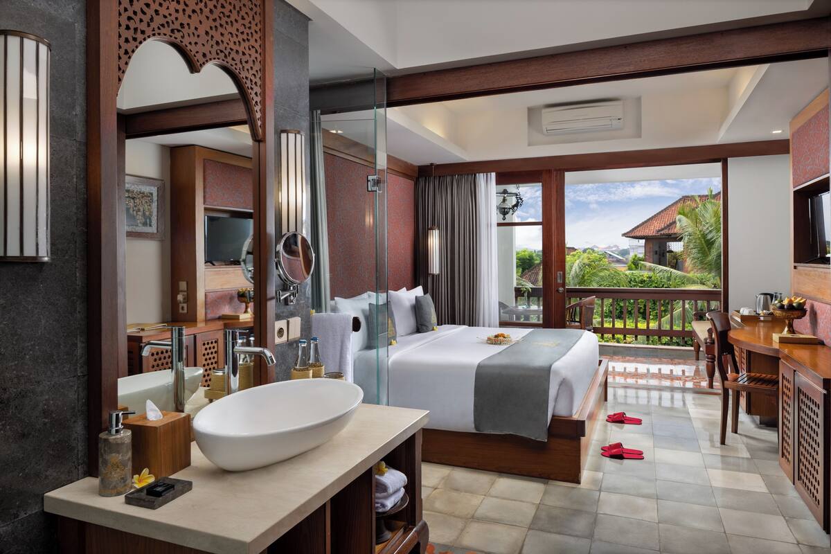 A modern hotel room with a large bed, a bathroom with a large sink and mirror, and a balcony with a view of the surrounding landscape.