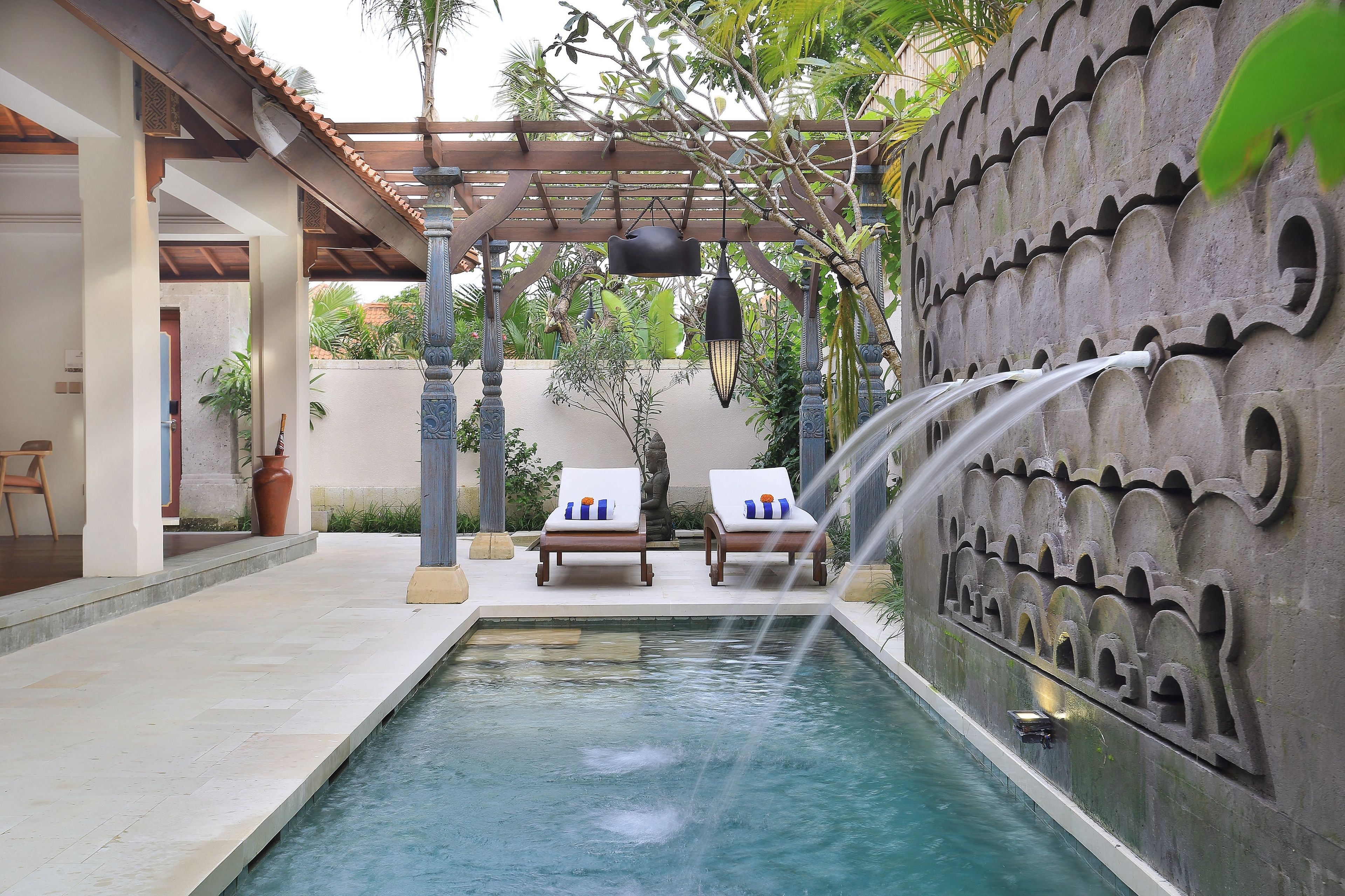 Área de piscina com um elemento aquático, duas espreguiçadeiras e um pergolado com luzes penduradas.