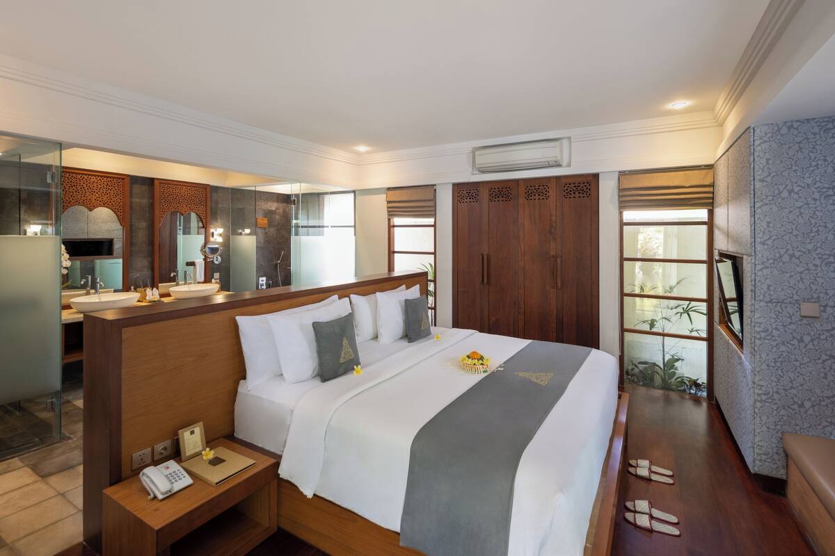 A hotel room with a large bed, a bedside table with a phone, a mirror, a vanity with a sink, and a view of a garden through a sliding door.