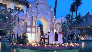 A serene evening setting with a water feature, a dining area with a table and chairs, and a decorative building with intricate designs. - The Alantara Sanur (Denpasar)