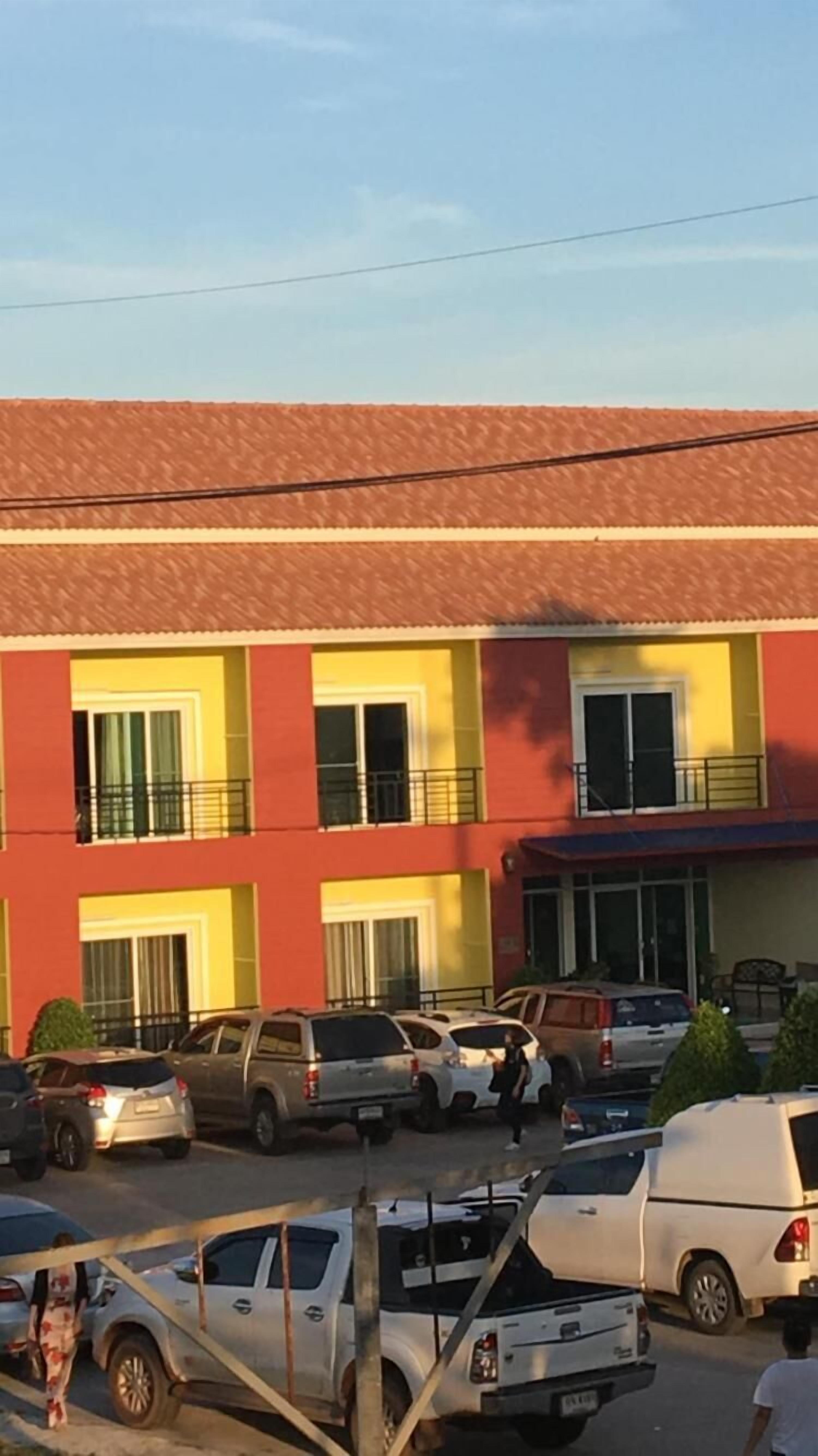 Un edificio de varios pisos con fachada roja y amarilla, varios balcones y un estacionamiento con distintos vehículos.