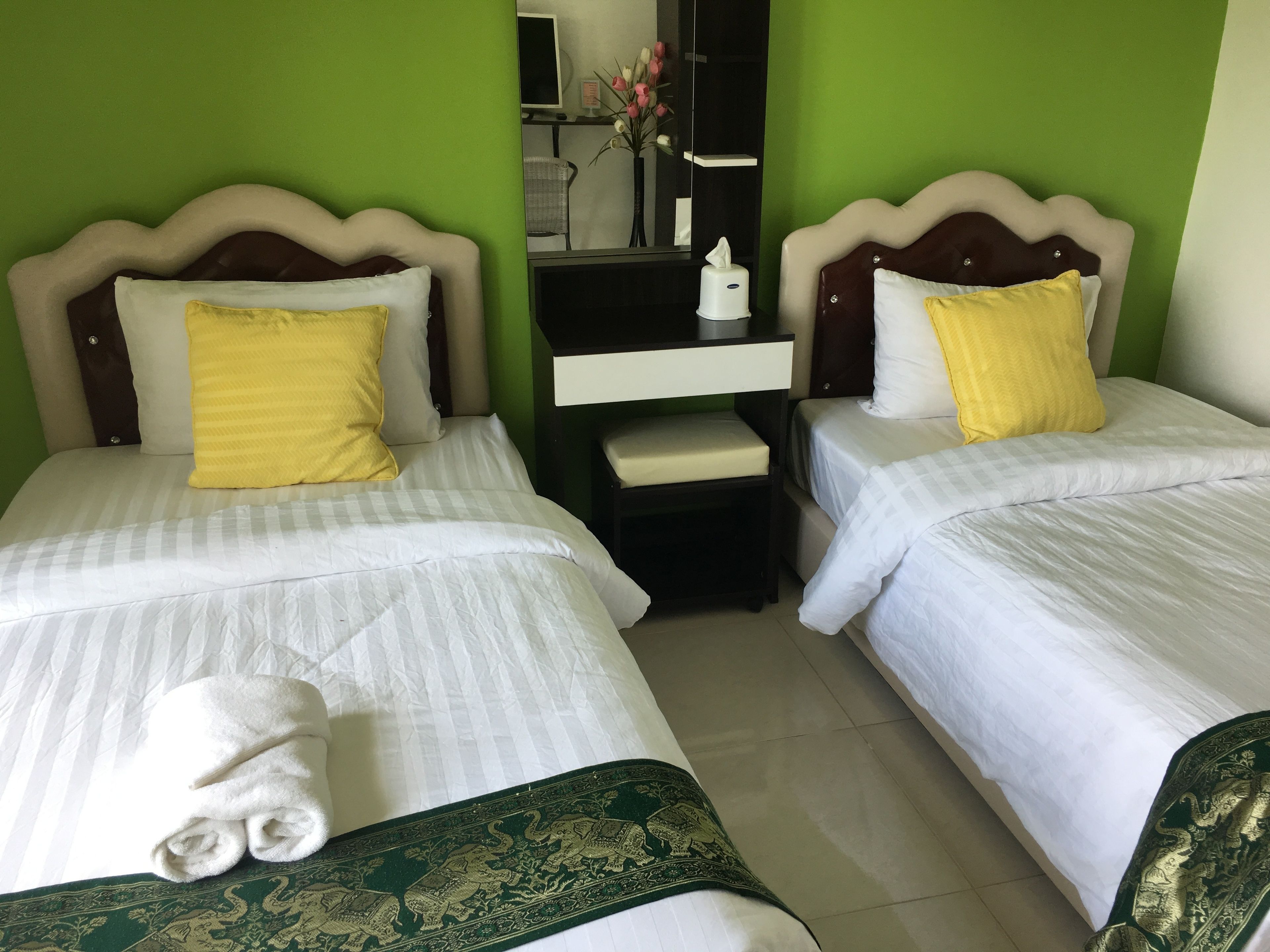 A hotel room with two beds, each with white bedding and yellow pillows. There is a small bedside table with a lamp and a tissue box. The room has green walls and a large mirror on the wall.