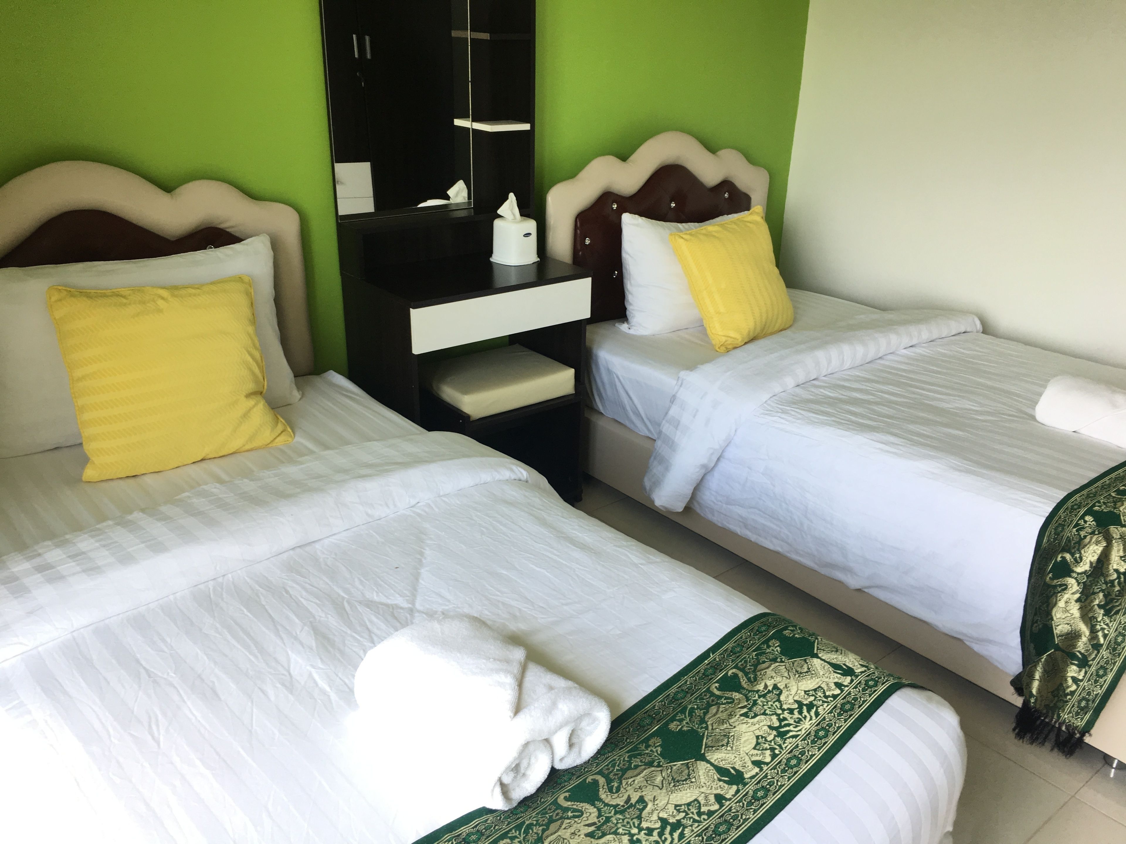 A hotel room with two single beds, each with a white bedspread and a yellow pillow. There is a nightstand with a mirror and a tissue box between the beds.
