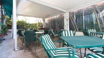 Un patio coperto con tavoli e sedie in metallo verde, piante in vaso e vista sul verde circostante.
