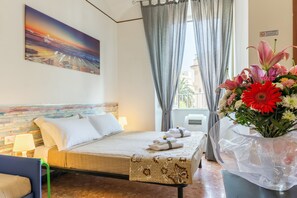 A hotel room with a bed, a chair, a painting on the wall, a window with curtains, and a vase of flowers. - Do Domus (Rome)