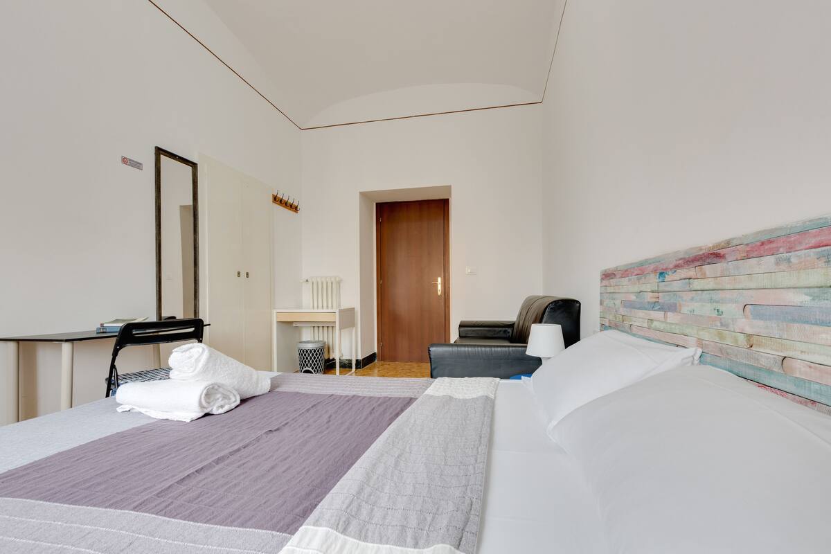 A hotel room with a bed, a desk, a chair, a mirror, and a door.