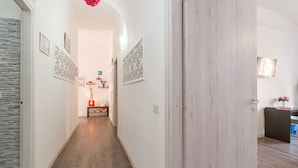 Un passadís amb terra de fusta, parets blanques i una porta a la dreta. Hi ha quadres amb marc a les parets i una taula petita amb un ram de flors a la distància.