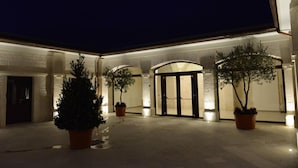 Un patio con una entrada central, árboles en macetas y paredes iluminadas durante la noche.
