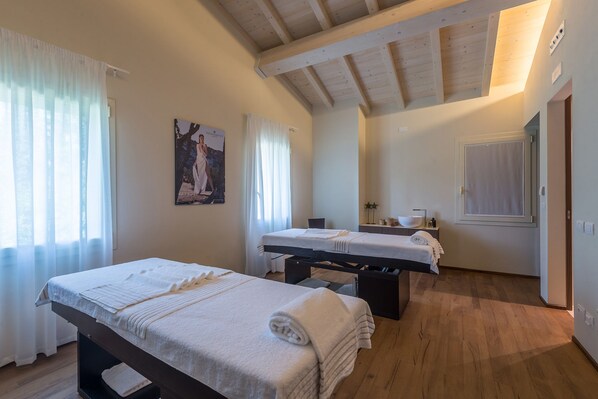 Una habitación con dos camas, techo de madera, un ventanal con cortinas, un cuadro en la pared y una mesita de noche con lámpara.