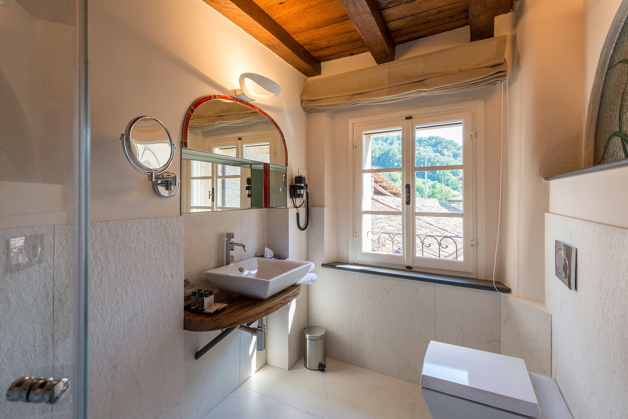 A modern bathroom with a large mirror, a sink, and a toilet. There is a window providing a view of the outside.