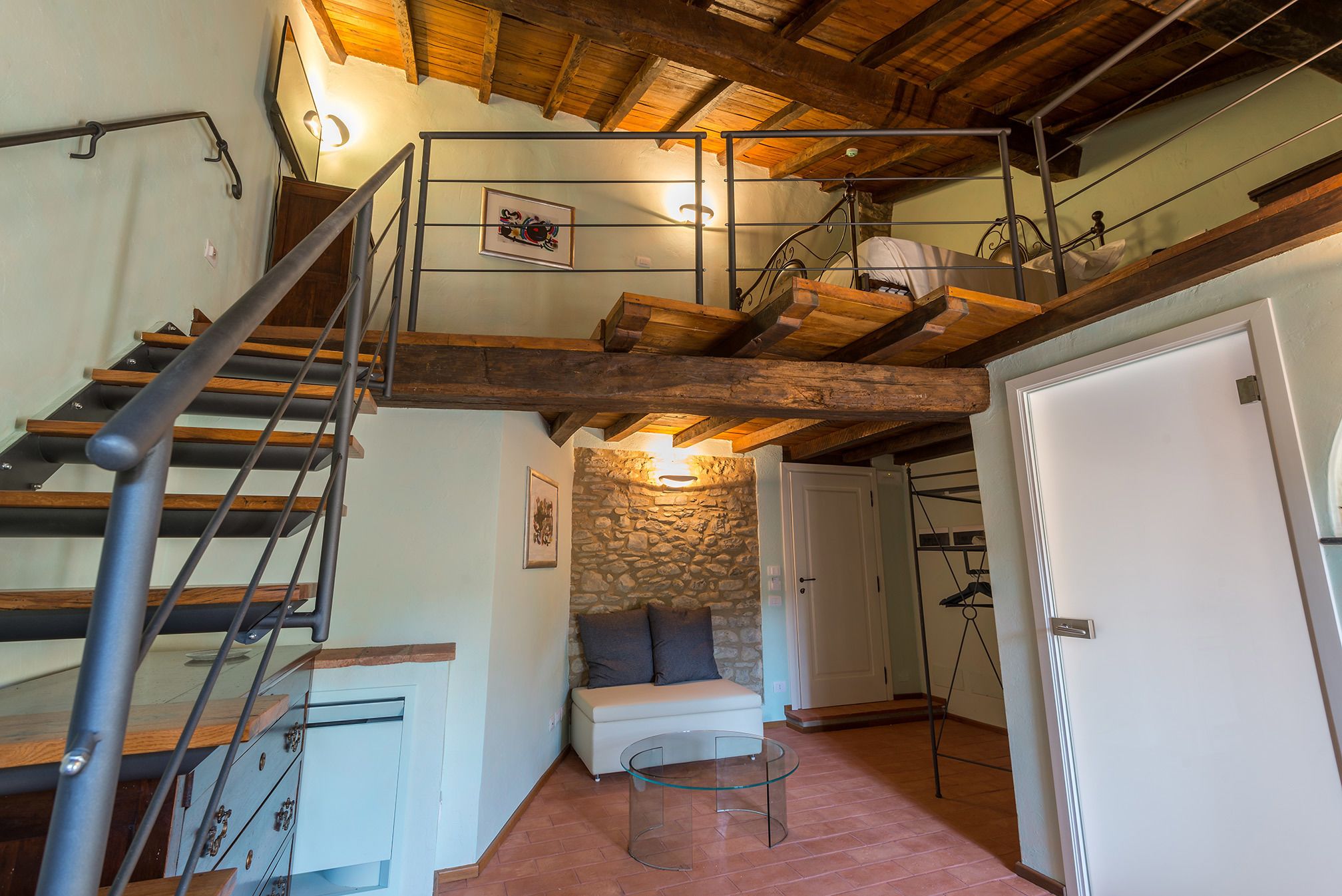 Una escalera de madera de varios niveles con barandillas de metal, un muro de piedra, un sofá blanco y una mesa de centro de vidrio en una habitación con suelo de baldosas.