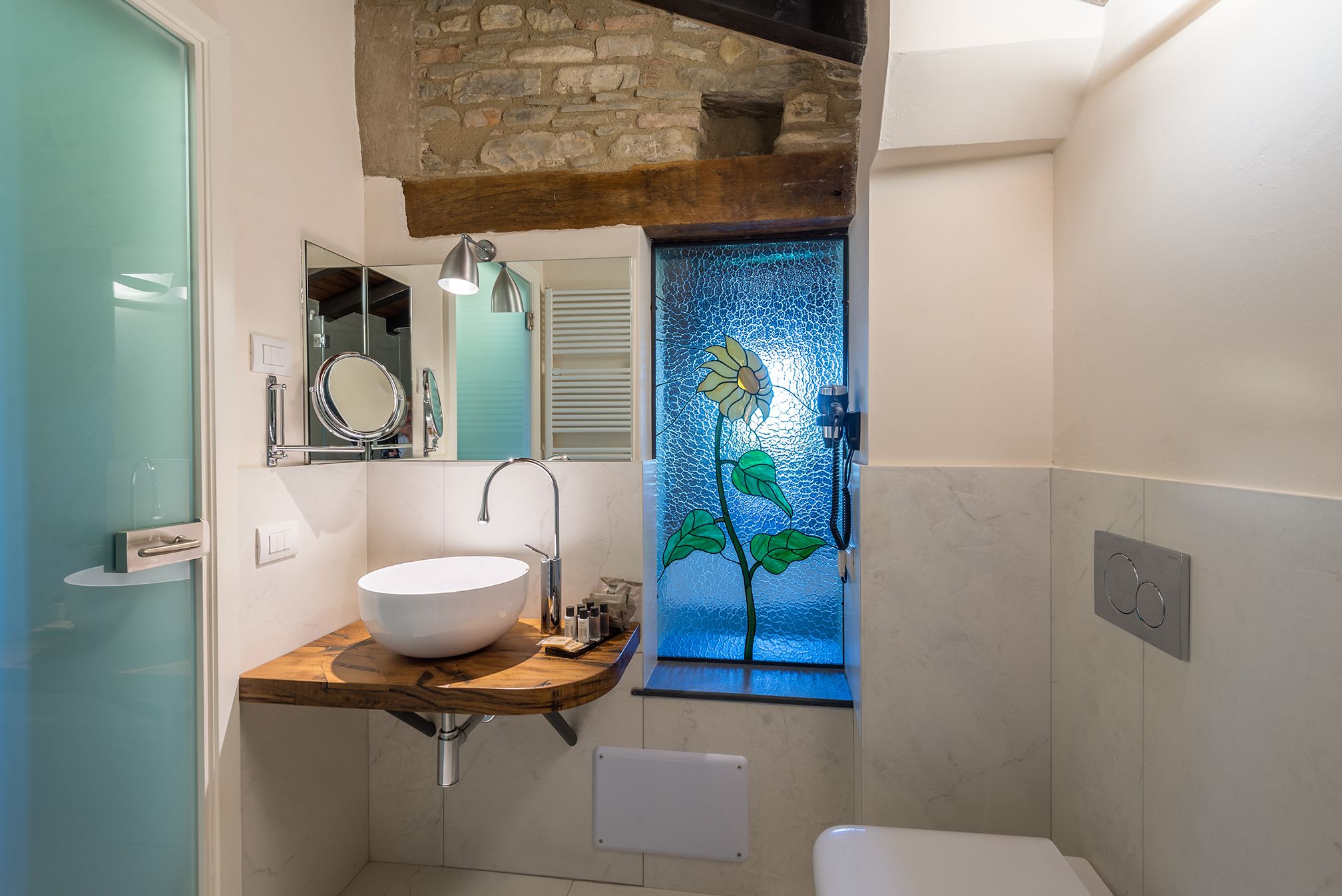 Ein Badezimmer mit einem großen weißen Waschbecken, einer Holzauslage, einem Spiegel, einer Glastür mit floralem Muster und einer Duschkabine.