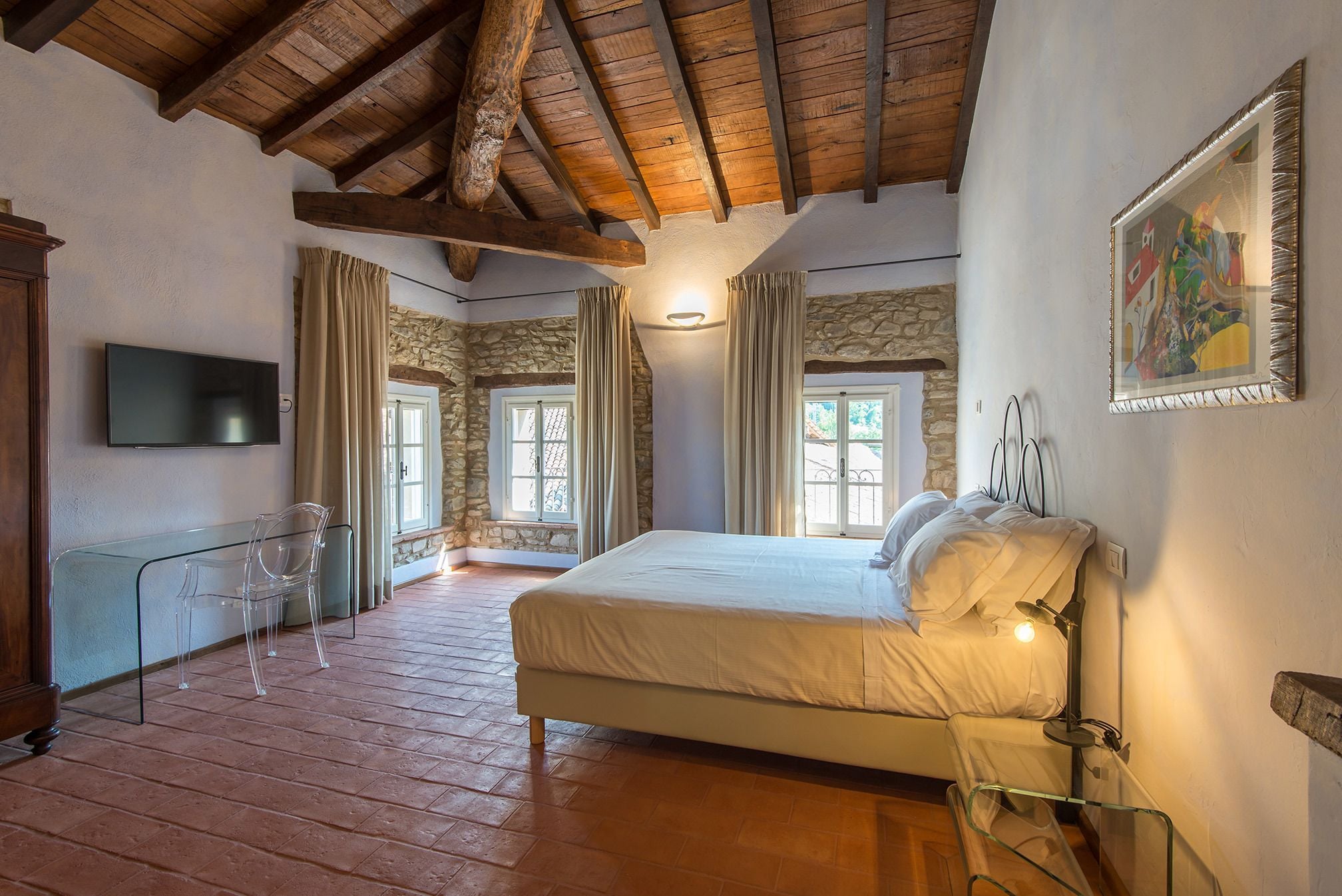 A bedroom with a stone wall, wooden beams, a bed, a TV, and a glass table.