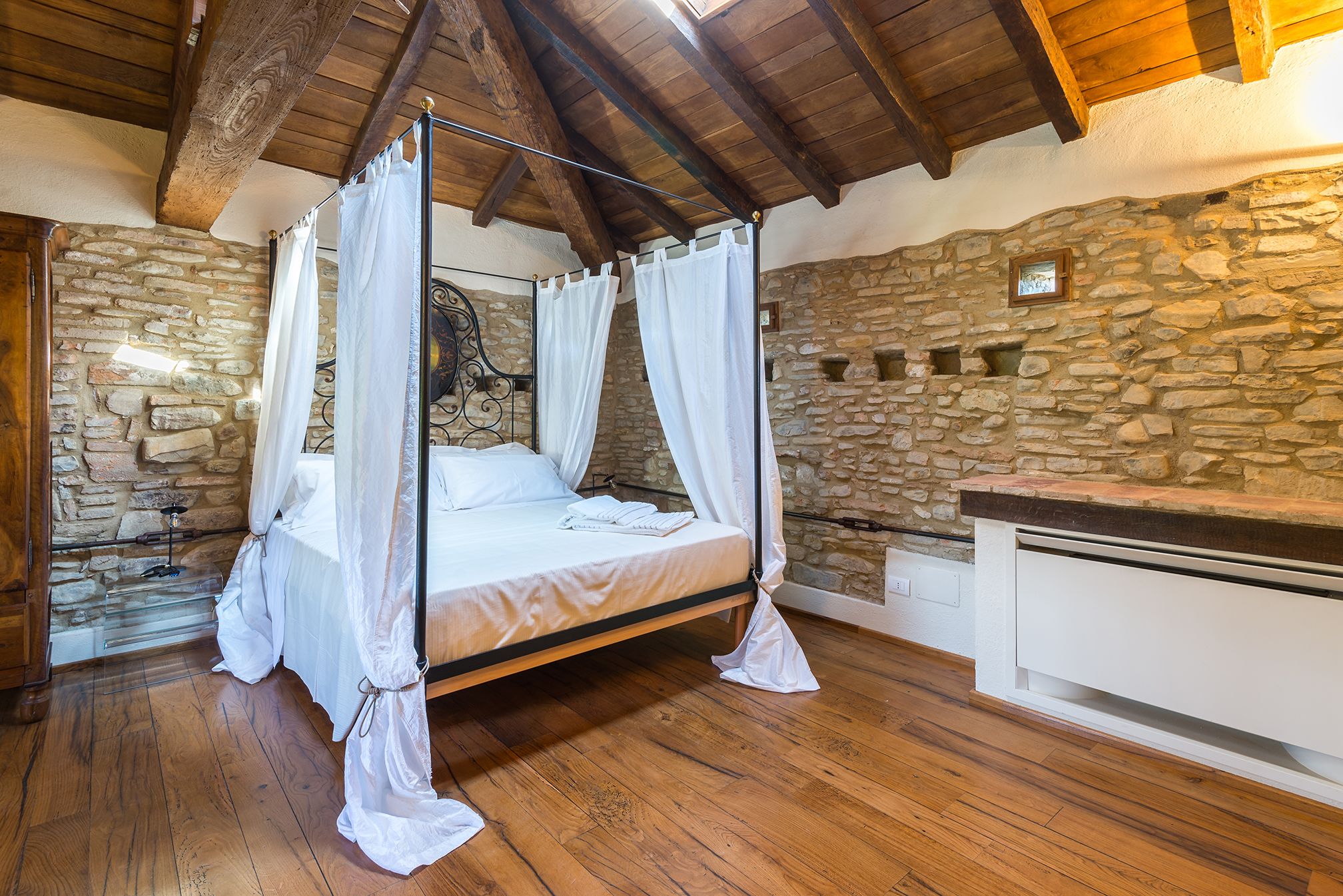 Ein Himmelbett mit weißen Leinen und einem Baldachin, vor einer Wand aus Stein und Holzbalken.