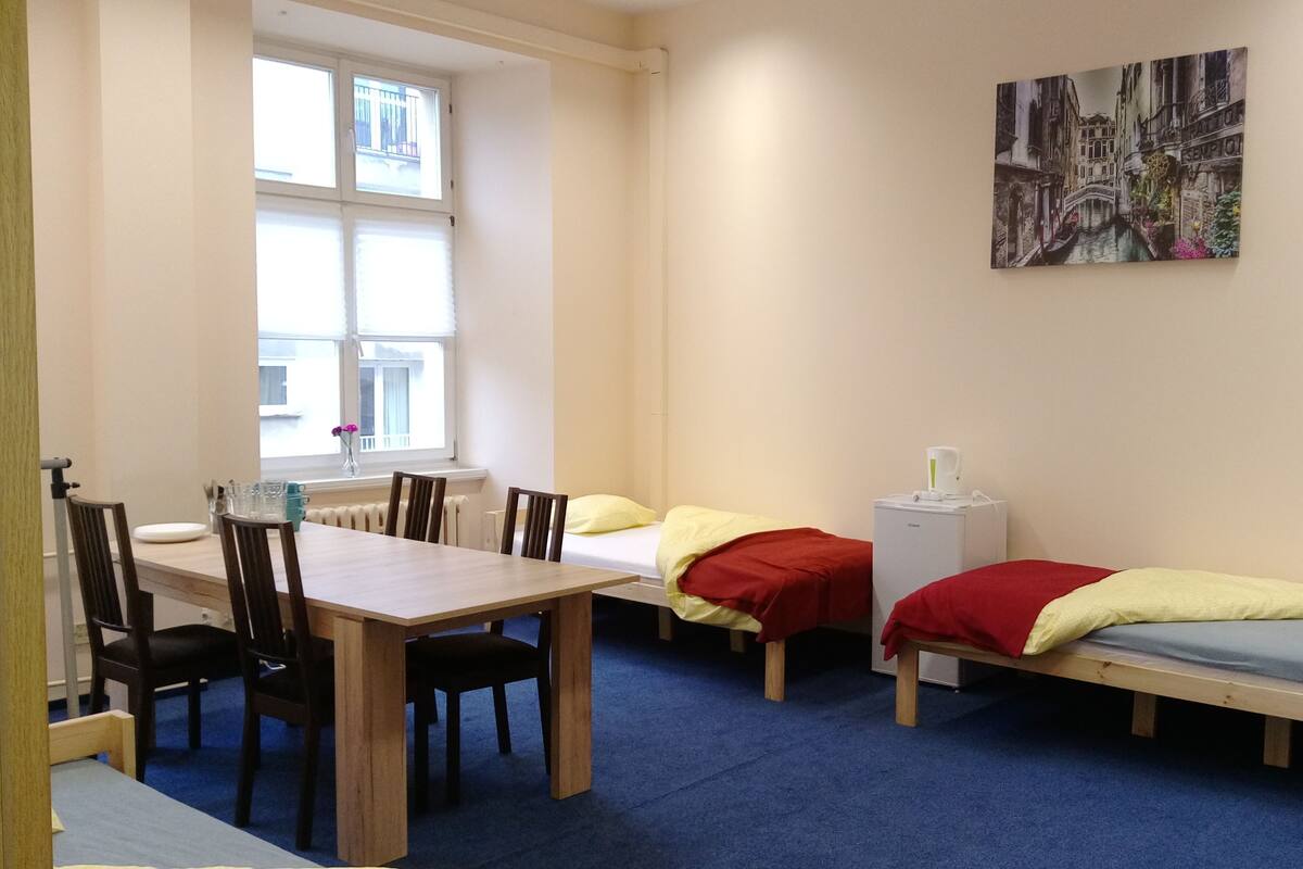 Una habitación con una mesa de madera, cuatro sillas, dos camas individuales con ropa de cama roja y amarilla, un radiador, una ventana con persianas y un cuadro en la pared.