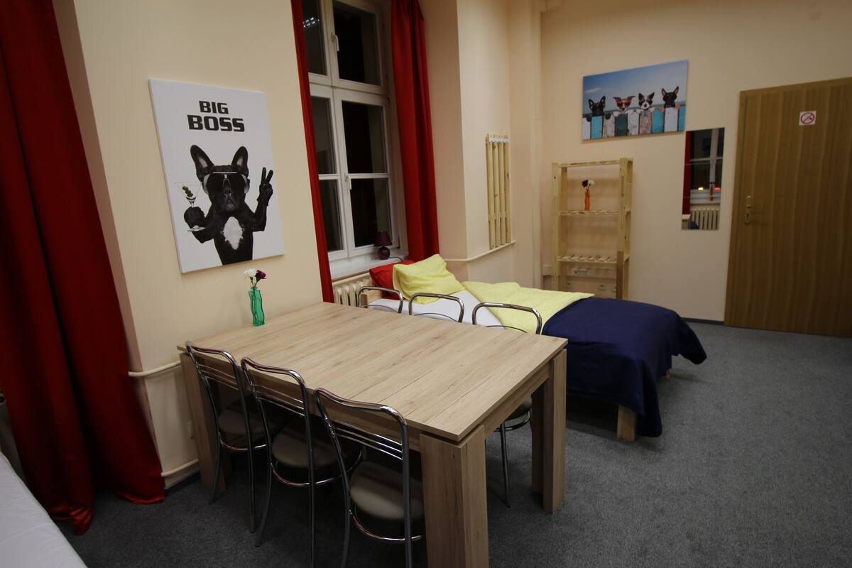 Una habitación con una mesa de madera y sillas, dos camas con ropa de cama azul y amarilla, un ventanal con cortinas rojas y un cartel de 'Big Boss' en la pared.