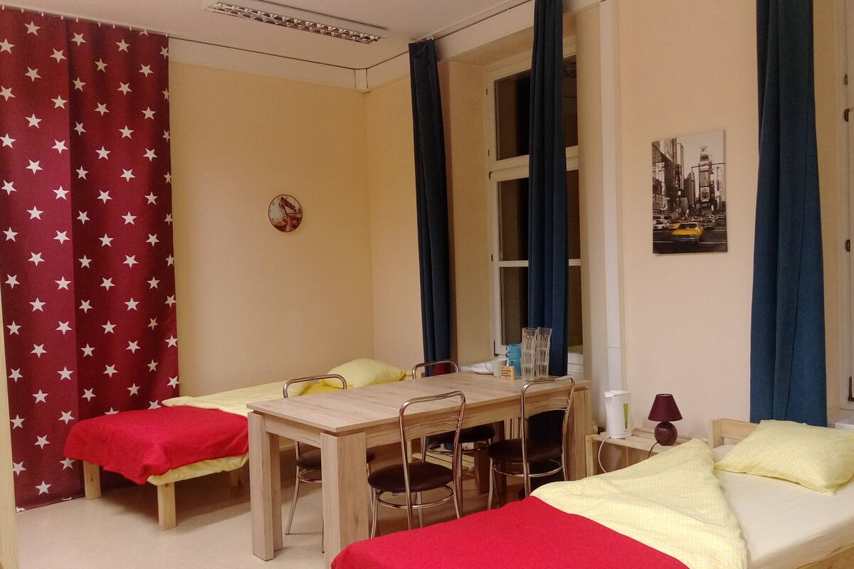 Una habitación pequeña y acogedora con dos camas individuales, una mesa de madera y sillas. Hay una ventana con cortinas, un cuadro colgado en la pared y una mesita con una lámpara y una taza.
